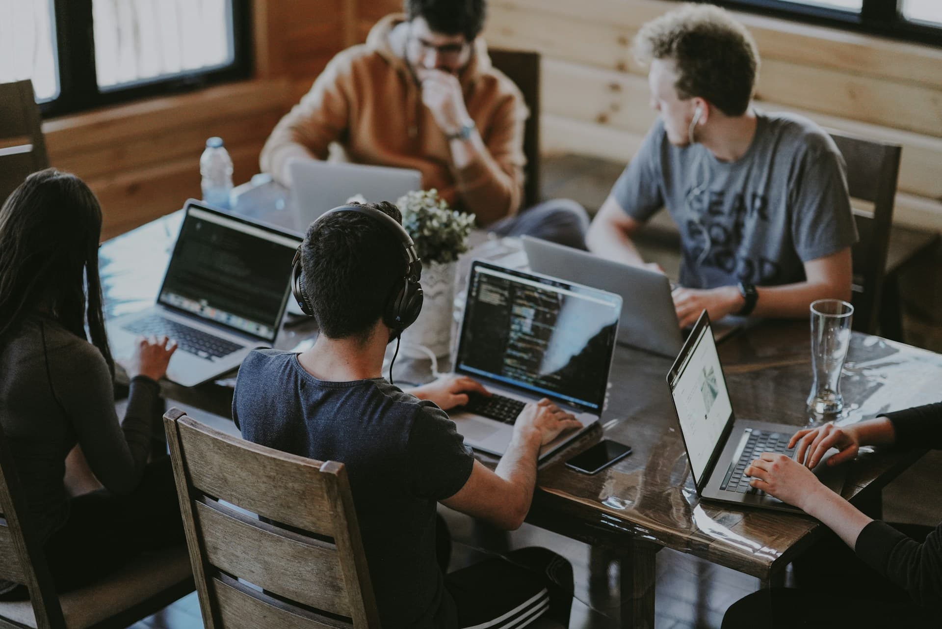 Fünf Menschen arbeiten an einem Holztisch an Laptops, auf zwei Bildschirmen ist Programmcode zu sehen.